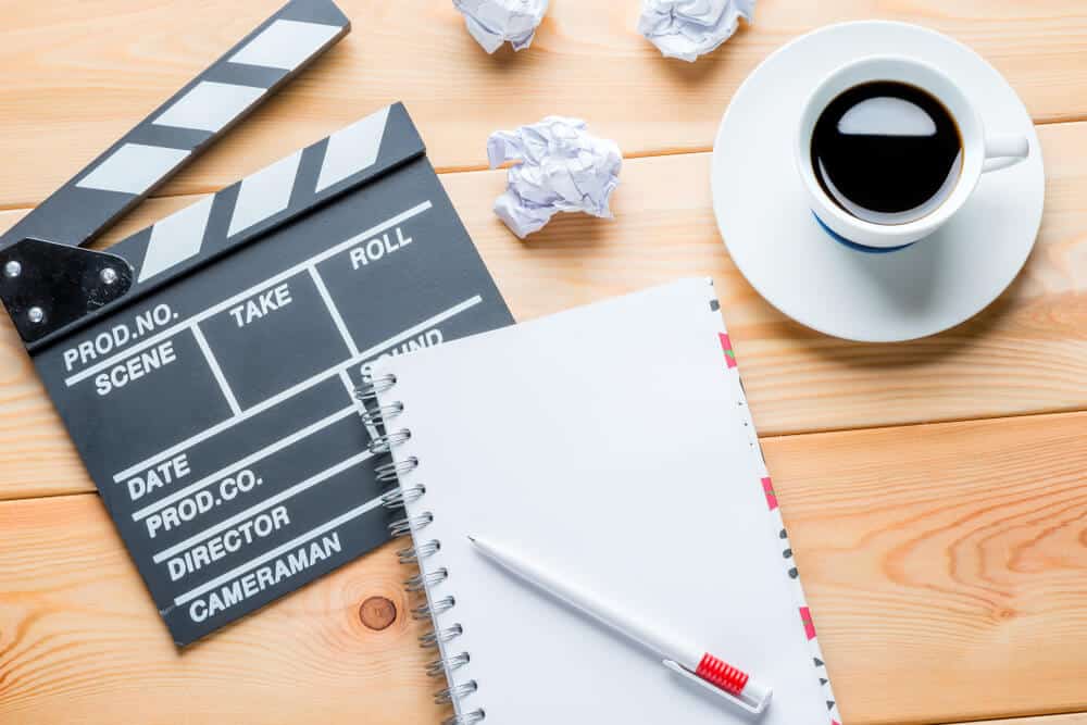 A clapperboard, notebook, pen, and a cup of coffee on a wooden table, with some crumpled paper scattered around while writing a script for a video