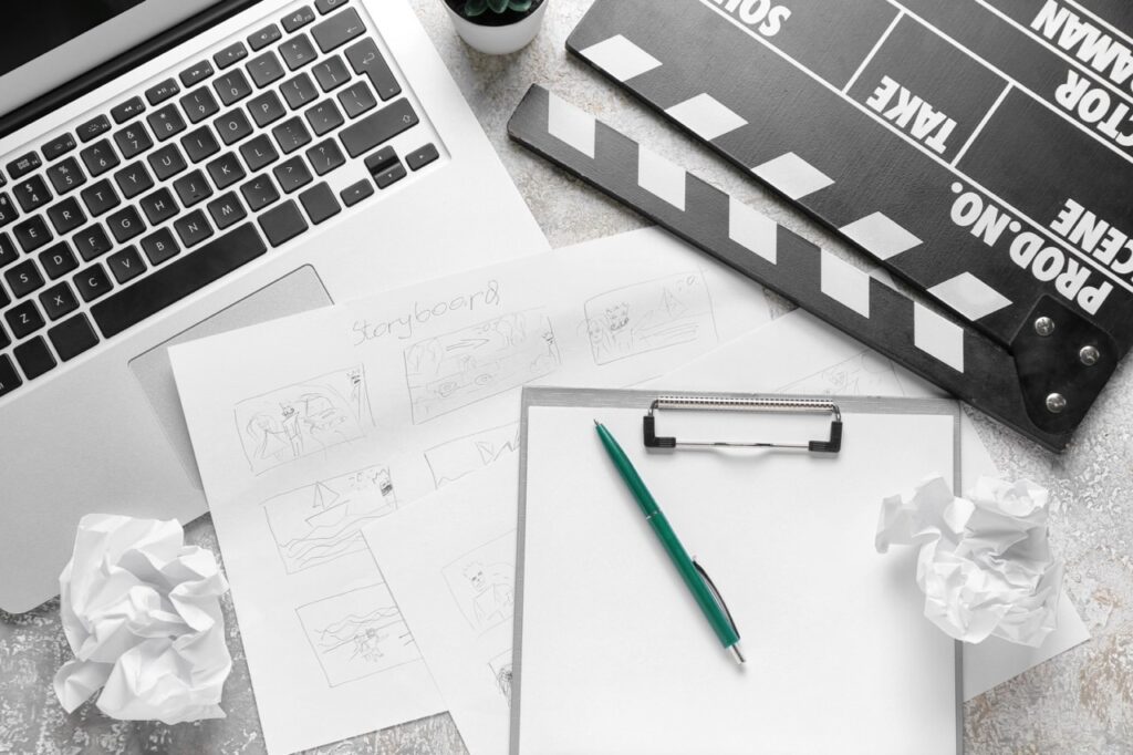 A laptop, storyboard sketches, a clipboard, and a clapperboard on a table, with crumpled paper nearby