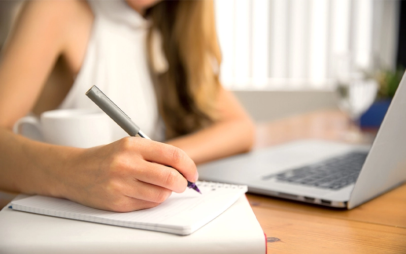 A person writing in a notebook with a pen while sitting at a desk with a laptop and a cup of coffee A person writing in a notebook with a pen while sitting at a desk with a laptop and a cup of coffee